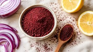 Small ceramic bowl heaped with deep crimson ground sumac on a cream linen cloth, a wooden spoon dusted with sumac