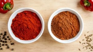 Overhead comparison of vibrant fresh red paprika and dull faded paprika in two matching white bowls on a light wood table, a few whole peppercorns and cumin seeds scattered