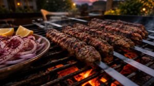 Charred kofta kebabs on flat metal skewers resting on a grill grate with flames softly glowing beneath, sumac dusted onions and lemon on the side